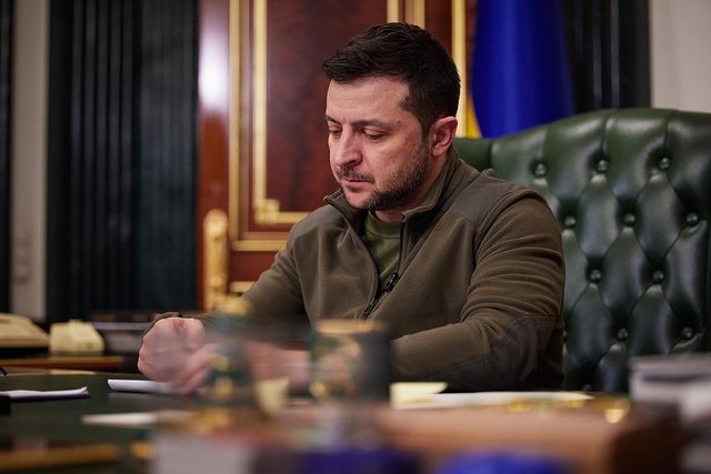 Volodymyr Zelenskyy in a brown jacket writing down a note on his desk.